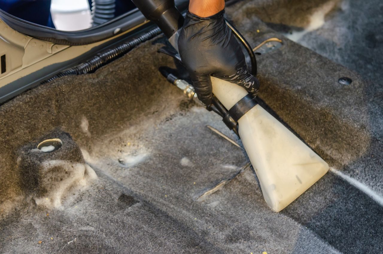 Home A hand in a glove using a vacuum to clean a car interior carpet, removing dirt effectively.